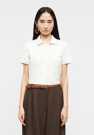 Ung kvinde med langt, glat, sort hår iført en hvid kortærmet skjorte med krave, brune plisserede bukser med høj talje, brun læderbælte og store gyldne øreringe.