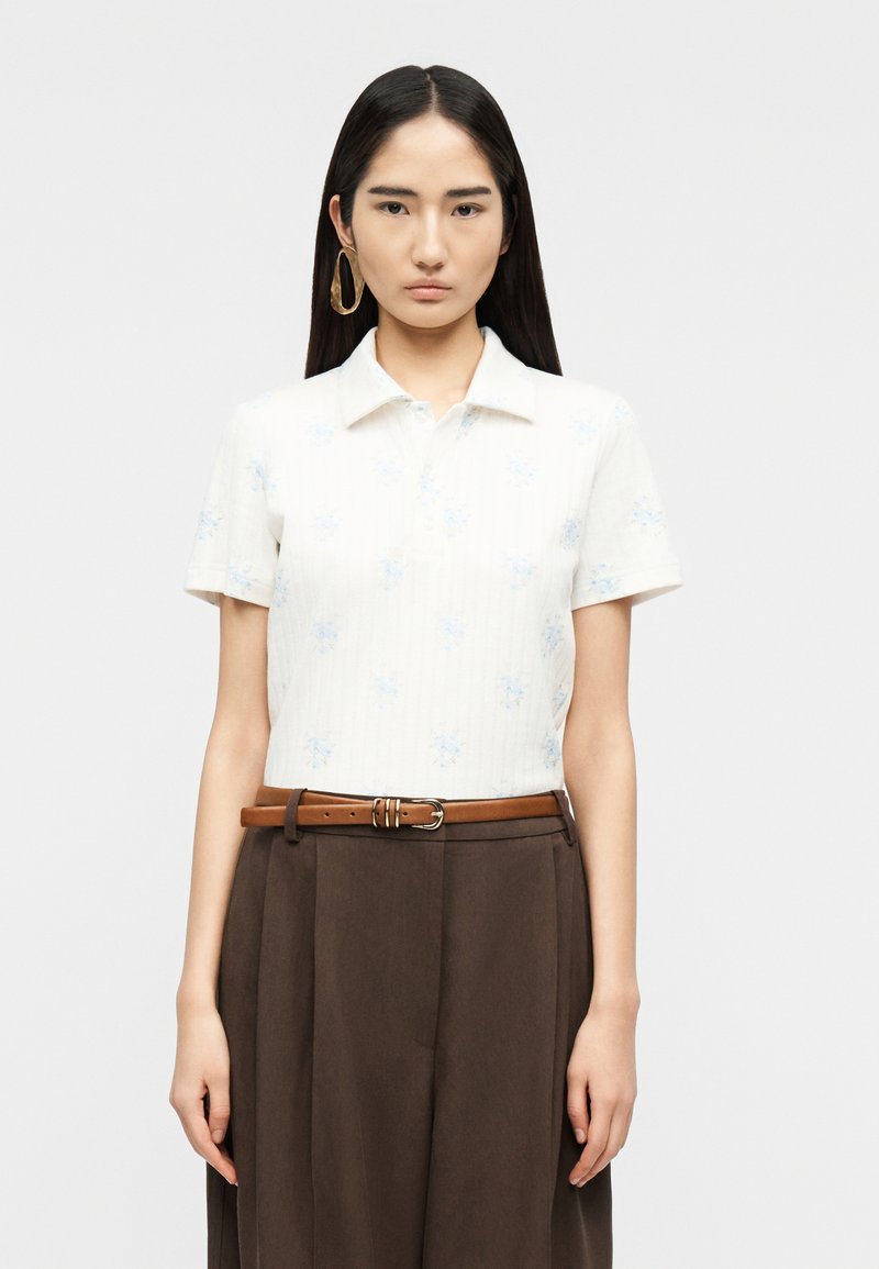 Jeune femme aux longs cheveux noirs et lisses portant une chemise blanche à manches courtes avec col, un pantalon plissé marron taille haute, une ceinture en cuir marron et une grande boucle d'oreille en or.