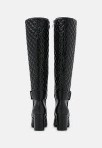 Botas negras hasta la rodilla con parte superior acolchada, cremallera lateral y tacones cuadrados, mostradas desde la parte posterior sobre un fondo liso.