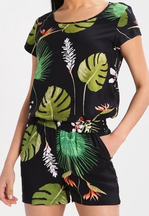 Vrouw draagt een zwart topje met korte mouwen en een korte broek met groene tropische bladeren en oranje bloemenprint, hand in de zak.