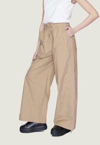 Pantaloni beige a gamba larga con vita regolabile, dotati di tasche laterali e righe verticali a contrasto. Indossati con scarpe platform nere.