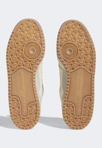 Suela de zapatos con una superficie de goma texturizada en color beige, que presenta patrones circulares y un logotipo en relieve. Bordes superiores lisos.