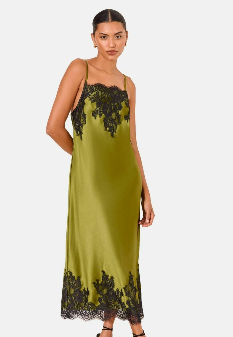 Woman wearing a sleeveless olive green satin dress with black lace trim on the neckline and hem, standing against a plain background.