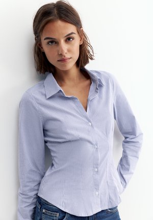 Ung kvinde med kort brunt hår iført en lyseblå stribet skjorte med knapper og blå jeans, står foran en hvid baggrund.