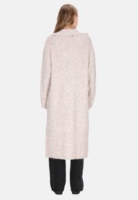 Persona con capelli lunghi che indossa un lungo cardigan beige lavorato a maglia, pantaloni neri e scarpe nere, in piedi di spalle contro uno sfondo neutro.