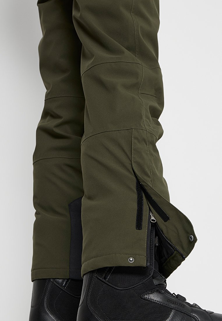 Nahaufnahme von olivgrünen Schneehosen mit seitlichem Reißverschluss und offenem Druckknopf über schwarzen Schneestiefeln.