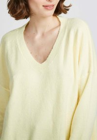 Pull en maille jaune clair, oversize, avec un décolleté en V. Texture douce, détails côtelés au niveau du décolleté et manchesraglan.