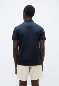 Μπλε σκούρο πόλο πουκάμισο από υφασμάτινη υφή, με κοντά μανίκια και κλασικό γιακά. Φοράται με ανοιχτόχρωμες βερμούδες, σε προβολή από πίσω.