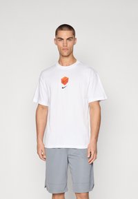 Biely bavlnený tričko s oranžovou grafikou v strede a čiernym logom Nike pod ním. V kombinácii so šedými športovými šortkami.