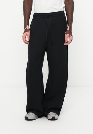 Black wide-leg sweatpants made of soft fabric with an elastic waistband and drawstring. Paired with gray athletic shoes.