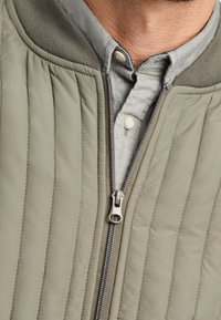 Giacca trapuntata verde oliva con zip frontale, colletto grigio e superficie testurizzata. Camicia a bottoni azzurra chiara visibile sotto.