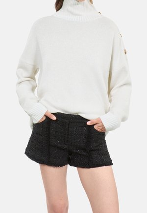Pull col roulé blanc avec boutons dorés sur l'épaule, associé à un short noir texturé taille haute, mains dans les poches.