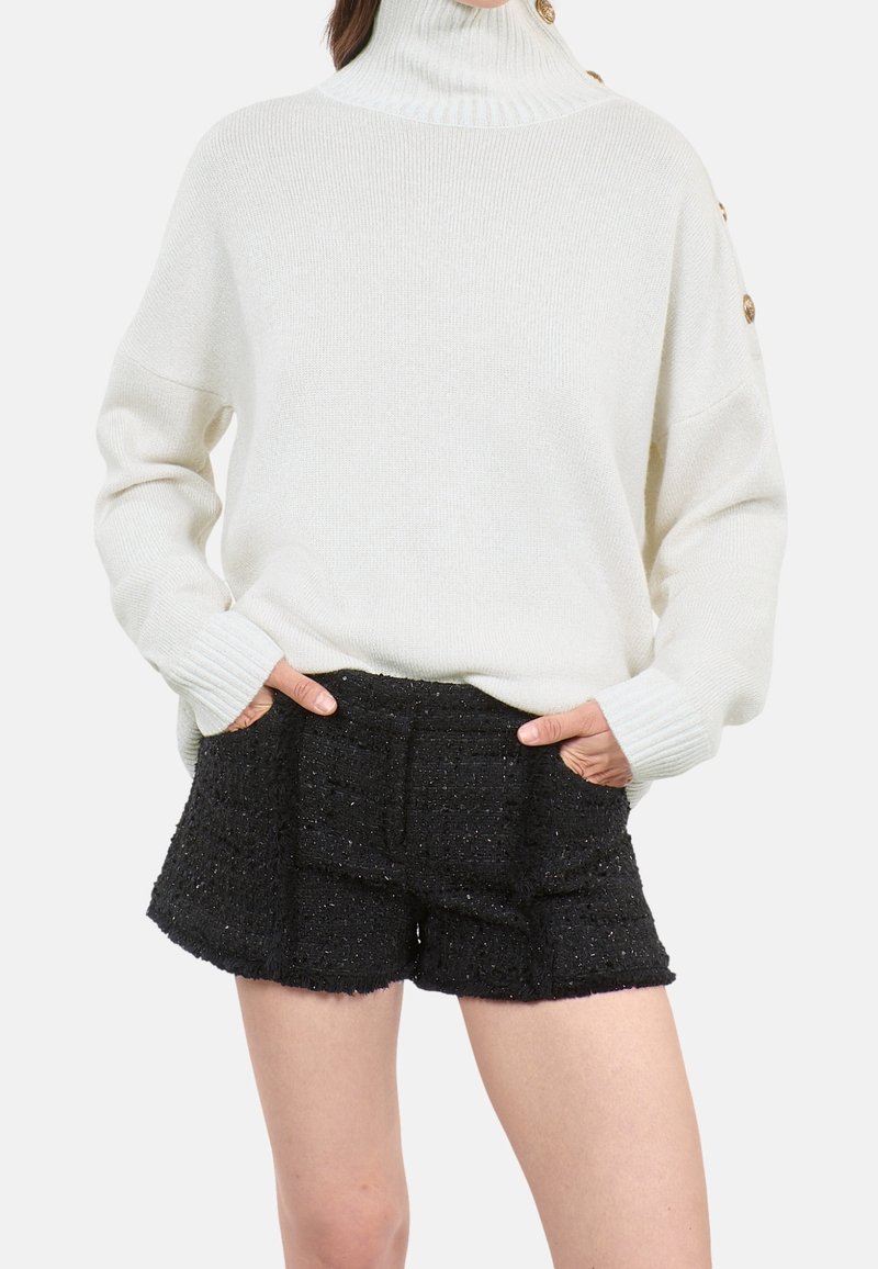 Pull col roulé blanc avec boutons dorés sur l'épaule, associé à un short noir texturé taille haute, mains dans les poches.