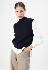 Frau mit zurückgegeltem Haar trägt eine marineblaue ärmellose Strickweste über einer weißen Bluse und hellbeigen Hosen, posiert mit einer Hand in der Tasche, neutraler Hintergrund.