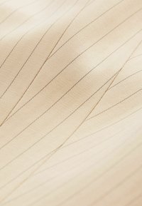 Tissu beige avec de fines coutures diagonales noires créant un motif géométrique sur une surface lisse et texturée.