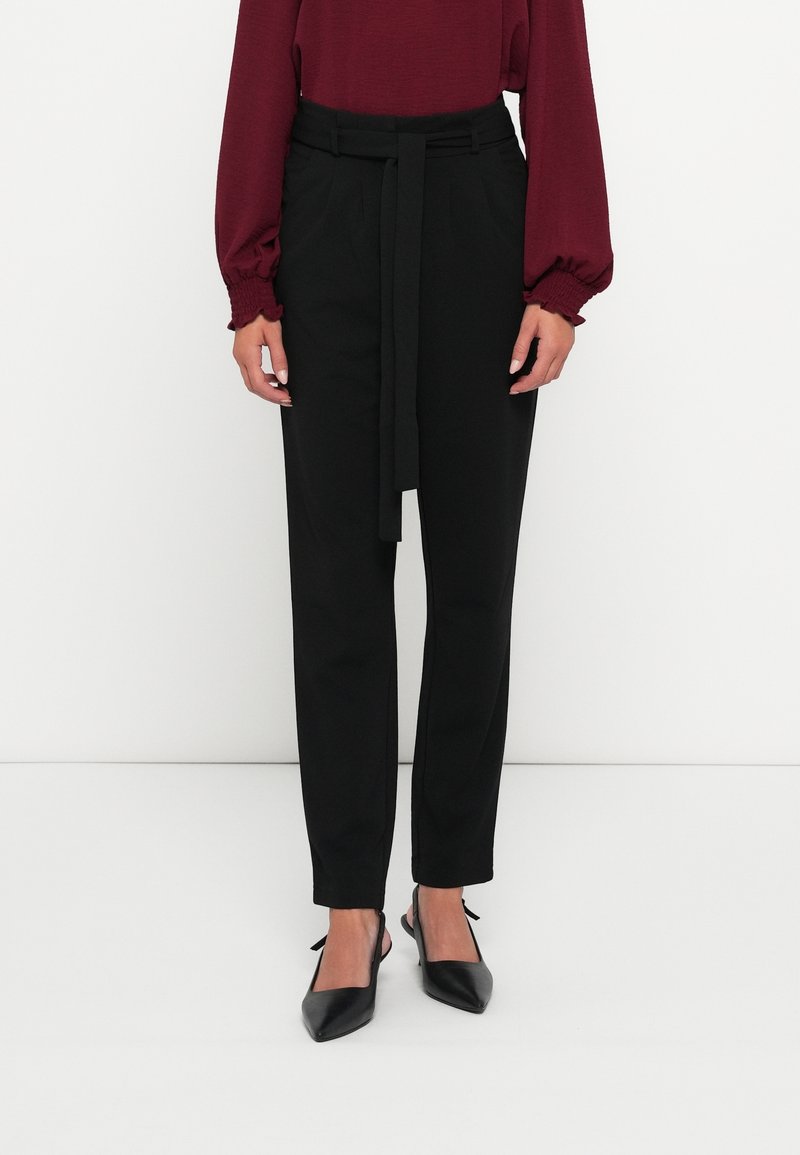 Femme portant un pantalon noir taille haute avec une ceinture, associé à un haut bordeaux à manches longues et des escarpins noirs à bout pointu.