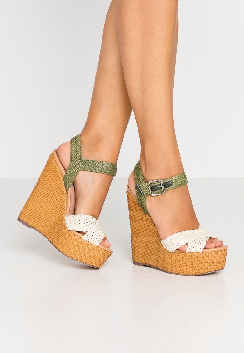Des jambes portant des sandales compensées tissées avec des semelles couleur cuir, des brides vertes à la cheville avec des boucles argentées, et des brides d'orteils tressées blanches sur un sol blanc.