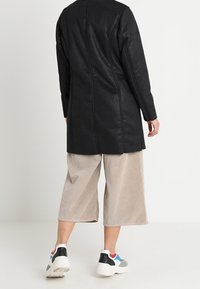 Černý kabát z umělé kůže se rovně střiženým střihem, s viditelnými švy. Modelka má na sobě světle béžové kalhoty s širokými nohavicemi a barevné sportovní boty.