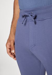 Lila Jogginghosen mit strukturierter Oberfläche, elastischem Bund und Kordelzug. Seiten Tasche auf der linken Seite sichtbar.