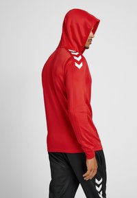 Rote Kapuzenjacke aus glattem Stoff mit weißen Zickzack-Akzenten auf den Schultern. Das Design ist tailliert mit langen Ärmeln.
