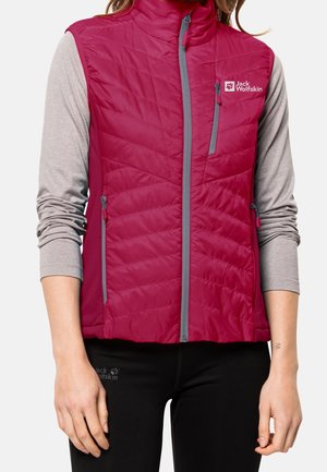 Kobieta ubrana w czerwoną, pikowaną kamizelkę bez rękawów z szarymi suwakami, na jasnoszarej koszuli z długim rękawem i czarnych spodniach.