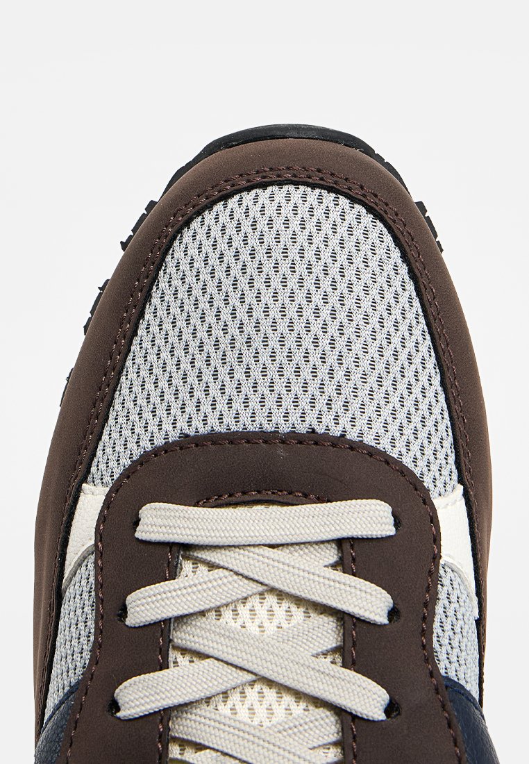 Zapatilla marrón y gris con parte superior de malla, cordones beige y suela de goma texturizada. Presenta un cuello acolchado y decoraciones de diseño sutiles.