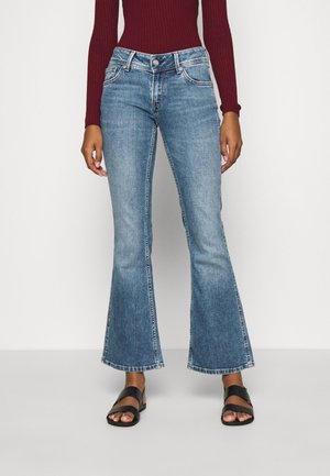 Kobieta w jasnoniebieskich jeansach z rozszerzanymi nogawkami, dopasowanym bordowym prążkowanym topie z długim rękawem oraz czarnych sandałach na odkryte palce, stojąca na białym tle.