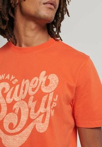 Orange bomulds t-shirt med rund hals og korte ærmer, med stor hvid tekst "Super Dry" med en tekstureret finish og subtilt mønster.
