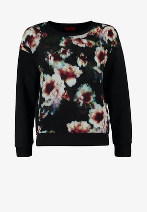 Femme portant un sweat-shirt noir à fleurs, un jean skinny bleu foncé, des bottines noires et un sac à bandoulière noir à franges.