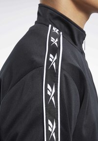 Černá atletická bunda s vysokým límcem, texturovanou látkou a kontrastními bílými pruhy s logem podél ramene.