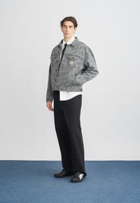 Chaqueta de mezclilla gris con una textura estampada, bolsillo en el pecho y botones, combinada con una camisa blanca y pantalones negros, sobre un fondo azul.