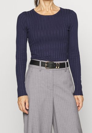 Femme portant un haut ajusté côtelé bleu marine à manches longues, un pantalon gris à fines rayures et une ceinture en cuir noire avec une boucle dorée.