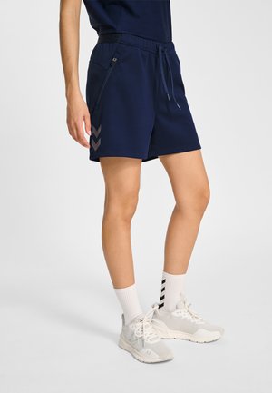 Osoba ubrana w granatowe sportowe spodenki z zamkami błyskawicznymi w kieszeniach, białe skarpetki za kostkę z czarnymi szewronami oraz białe tenisówki na jednolitym białym tle.