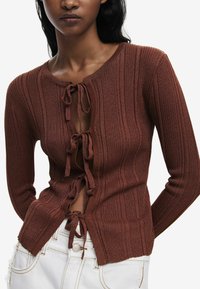 Brun ribbet cardigan med dyb V-hals, der har snører ned foran. Materialet ser ud til at være strikket med en blød tekstur, båret over hvide bukser.