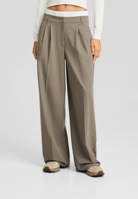 Pantalones de pierna ancha en una suave tela gris-marrón, con un diseño plisado y una cinturilla elástica blanca. Combinados con zapatillas neutras.