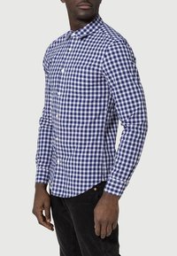 Blå och vit rutad skjorta med lång ärm, spetsig krage, knapprad framtill och böjd nederkant, i kombination med mörka byxor.