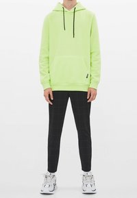 Limegrön hoodie med en framficka och svarta dragsnören, kombinerad med svarta rutiga tapered byxor och vita sportskor.