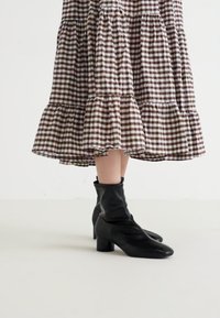 Bottines noires en matériau lisse avec un bout arrondi et un talon bloc, associées à une robe à volants à carreaux marron et blanc.