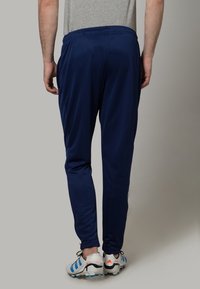 Námořnické modré sportovní joggery s tapered střihem, elastickým pasem a jemným leskem; nošeny s bílými a modrými sportovními botami.