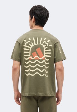 Homme portant un t-shirt vert olive avec un motif stylisé de coucher de soleil, nuages et vague dans le dos, debout dos à la caméra.