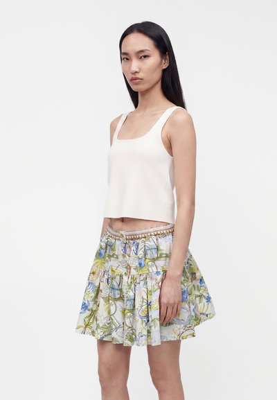 Νεαρή γυναίκα που φοράει ένα άσπρο κοντό τοπ χωρίς μανίκια και μια χρωματιστή πλισέ φούστα με λουλουδάτο σχέδιο, στέκεται μπροστά σε ένα απλό φόντο.