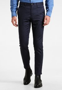 Man som bär slim-fit mörkblå byxor, ljusblå skjorta och svarta läderankelstövlar som går på vit bakgrund.