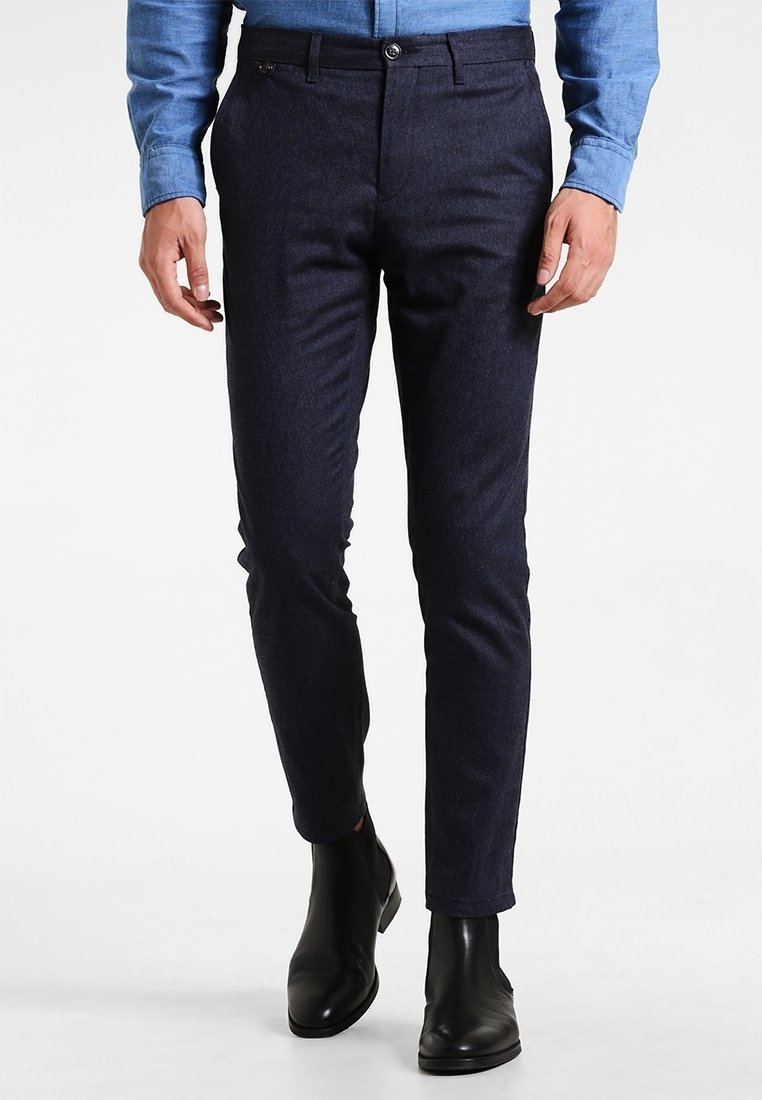 Man som bär slim-fit mörkblå byxor, ljusblå skjorta och svarta läderankelstövlar som går på vit bakgrund.