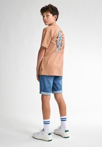 Camiseta color marrón claro con un gráfico colorido en la parte posterior, combinada con pantalones cortos de denim arremangados y zapatillas blancas con acentos verdes y calcetines a rayas azules.
