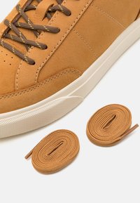 Baskets en daim beige avec une semelle en caoutchouc crème, dotées de lacets marron avec des accents jaunes. Deux lacets beige supplémentaires sont présentés à côté de la chaussure.
