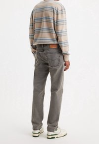 Persona vestida con jeans rectos grises, camiseta de manga larga a rayas beige y azul, y zapatillas blancas, de pie contra un fondo blanco.
