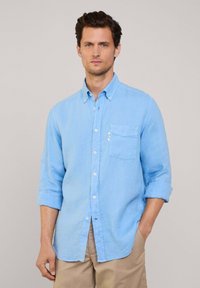 Camicia button-up azzurra in tessuto di lino con maniche lunghe arrotolate, dotata di taschino sul petto e bottoni bianchi.
