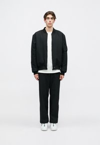 Melnā bumbiera jaka ar gludu tekstūru, kārtota virs baltas krekla, kombinēta ar melnām biksēm un baltām sporta kedām. Modelis stāv taisni.