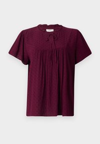 Blouse bordeaux à manches courtes avec motif à points texturés, encolure ronde à volants et détail noué à l'avant sur fond uni.