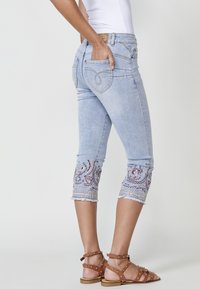 Light blue cropped jeans feature embroidered paisley designs and frayed hems. Back pockets have decorative stitching. Paired with brown strappy sandals.